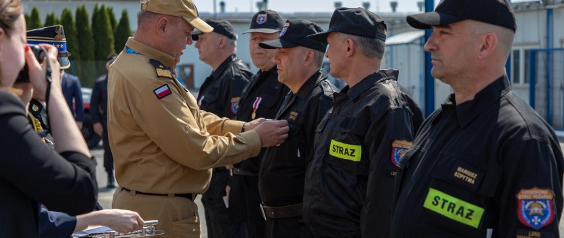 Zdjęcie wykonane na zewnątrz, podczas uroczystej zbiórki. Oficer Państwowej Straży Pożarnej w stopniu generała przypina odznaczenia strażaków stojącym w szeregu.