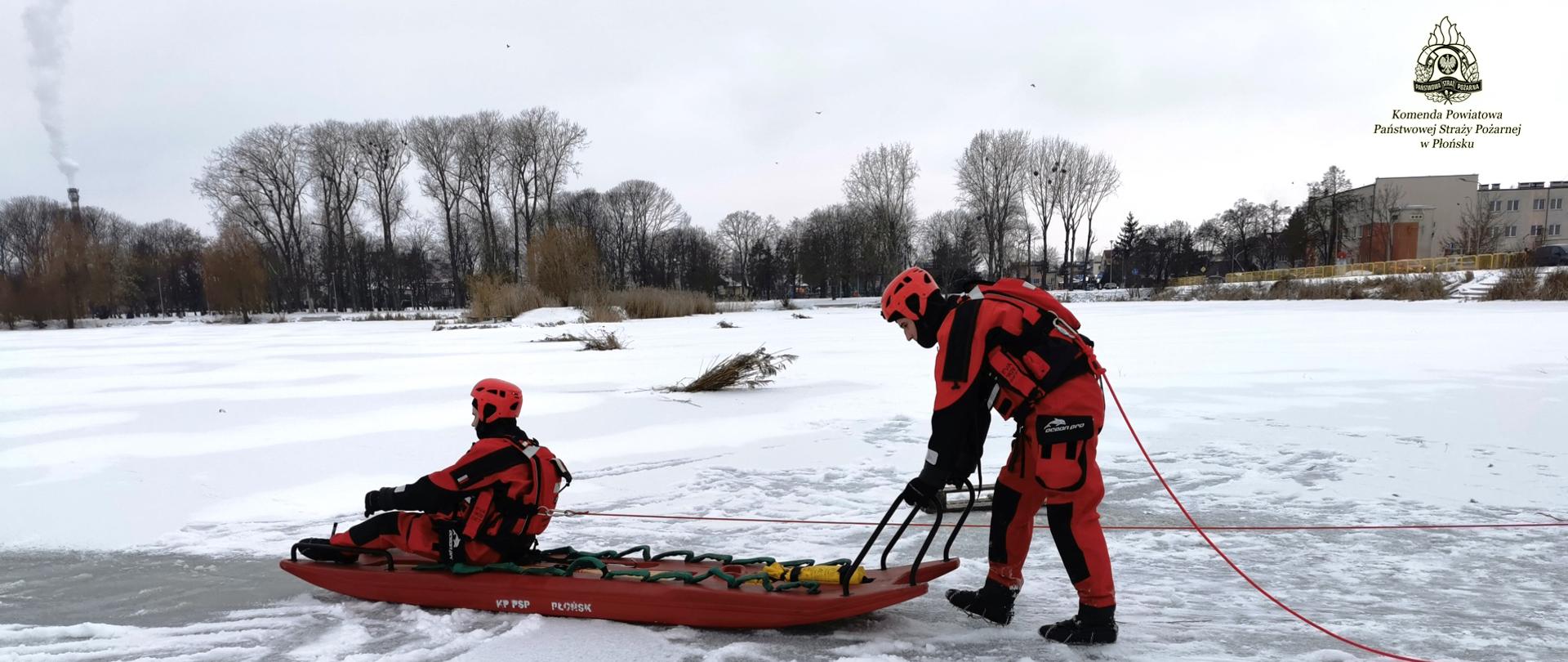 Dwóch strażaków ubranych w ubrania do ratownictwa lodowego. Pierwszy siedzi na saniach lodowych, drugi pcha sanie. Zdarzenie ma miejsce na zamarzniętym zbiorniku wodnym.