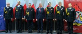 Sześciu mężczyzn w galowych mundurach strażackich. Po środku nich starszy mężczyzna w garniturze. Stoją w szeregu na tle biało-czerwonych flag 