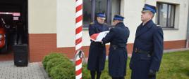 Narodowe Święto Niepodległości. Plac przed Komendą Powiatową PSP w Rawiczu. Na tle budynku komendy, przed biało-czerwonym masztem stoi poczet flagowy. Dowódca pocztu wraz z flagowym przygotowują flagę do zamocowania. Asystujący stoi w postawie zasadniczej.