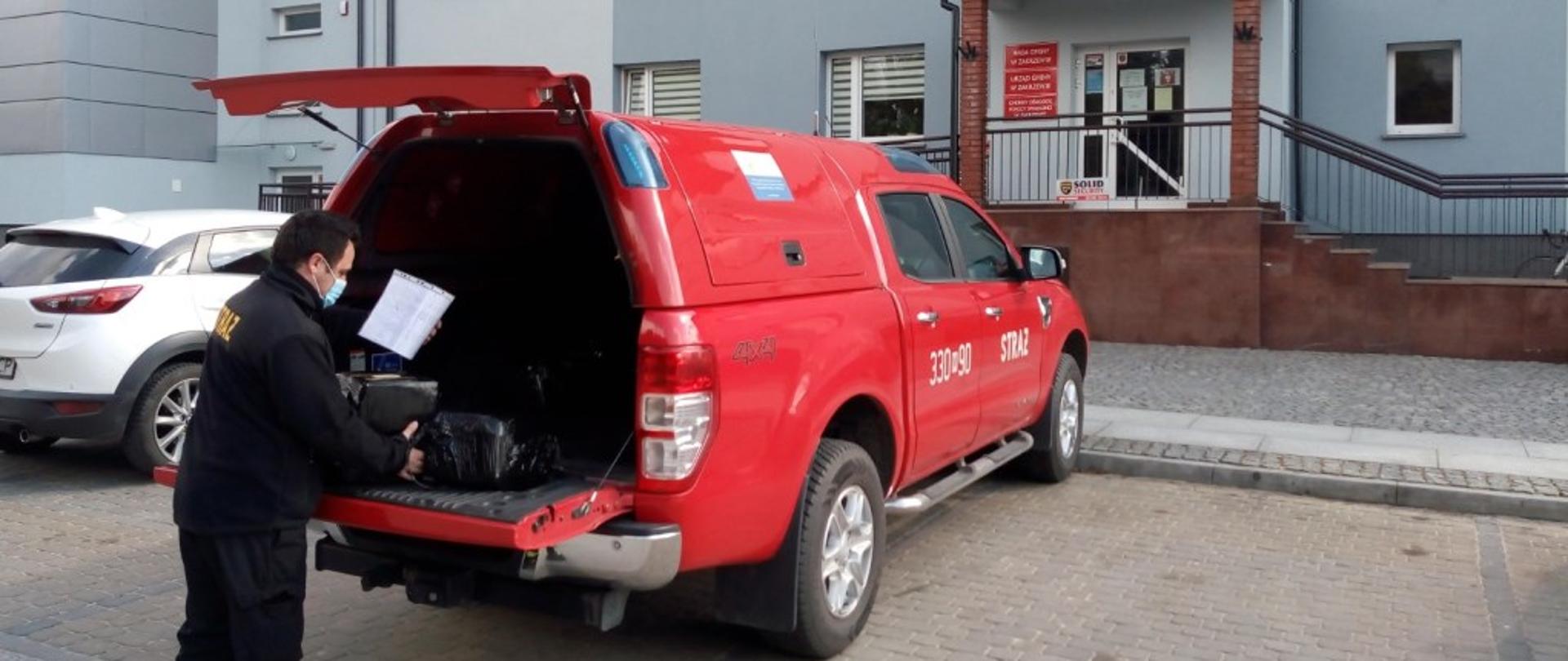 Zdjęcie przedstawia pojazd specjalny terenowy typu pick-up stojący przed budynkiem urzędu gminy. Pojazd w kolorze czerwonym ze srebrnymi elementami zabudowy. Tylna klapa pojazdu jest otwarta. Przy niej stoi strażak ubrany w ubranie koszarowe w kolorze czarnym z jaskrawymi elementami odblaskowymi. W lewej ręce trzyma kartkę. Prawą ręką przesuwa opakowania z ulotkami leżące na otwartej klapie bagażnika. Na twarzy ma założoną maseczkę.