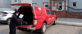 Zdjęcie przedstawia pojazd specjalny terenowy typu pick-up stojący przed budynkiem urzędu gminy. Pojazd w kolorze czerwonym ze srebrnymi elementami zabudowy. Tylna klapa pojazdu jest otwarta. Przy niej stoi strażak ubrany w ubranie koszarowe w kolorze czarnym z jaskrawymi elementami odblaskowymi. W lewej ręce trzyma kartkę. Prawą ręką przesuwa opakowania z ulotkami leżące na otwartej klapie bagażnika. Na twarzy ma założoną maseczkę.