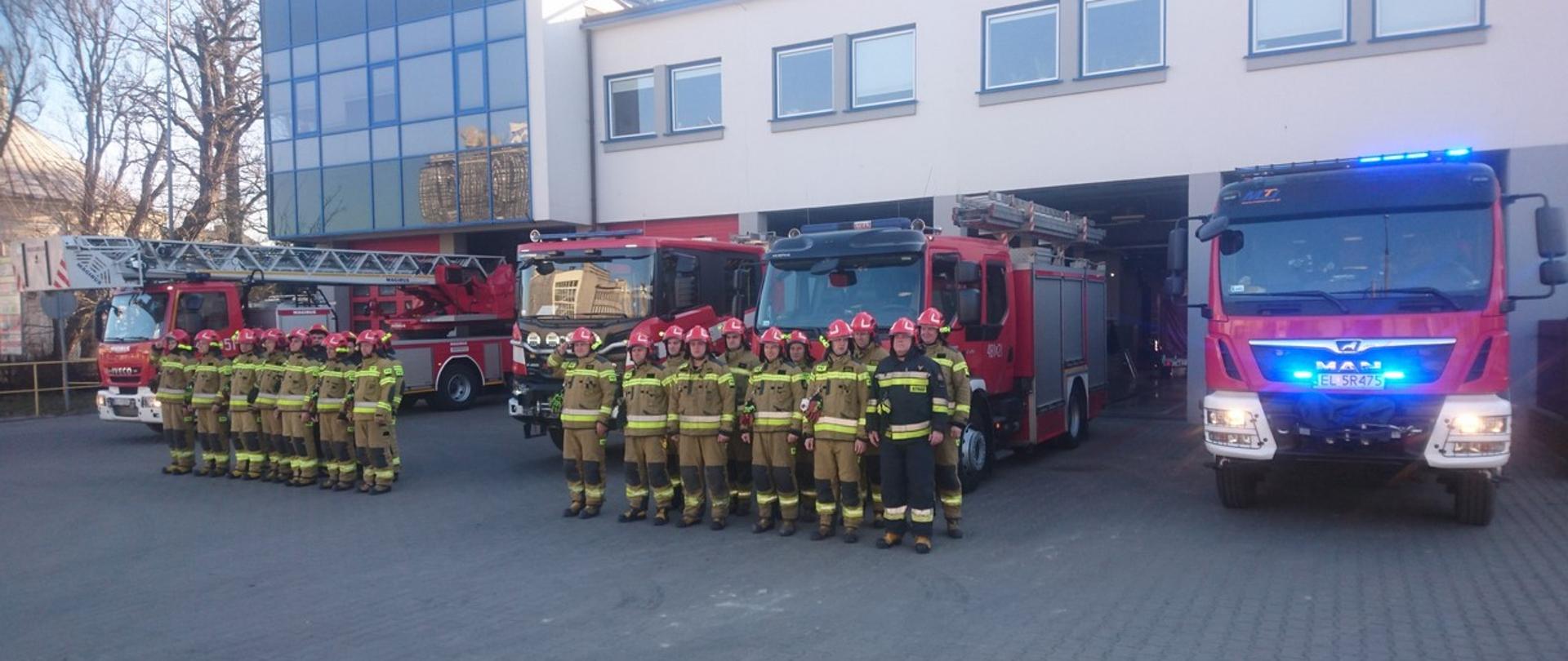 Zdjęcie przedstawia dwie zmiany służbowe z JRG w Pabianicach. Strażacy oddają cześć poległym ukraińskim strażakom. Bezpośrednio za strażakami ustawione są samochody strażackie, od lewej: samochód z drabiną mechaniczną, ciężki samochód ratowniczo gaśniczy, średni samochód ratowniczo gaśniczy i ostatni po prawej to średni samochód ratowniczo-gaśniczy, W tle budynek Komendy Powiatowej PSP w Pabianiach.
