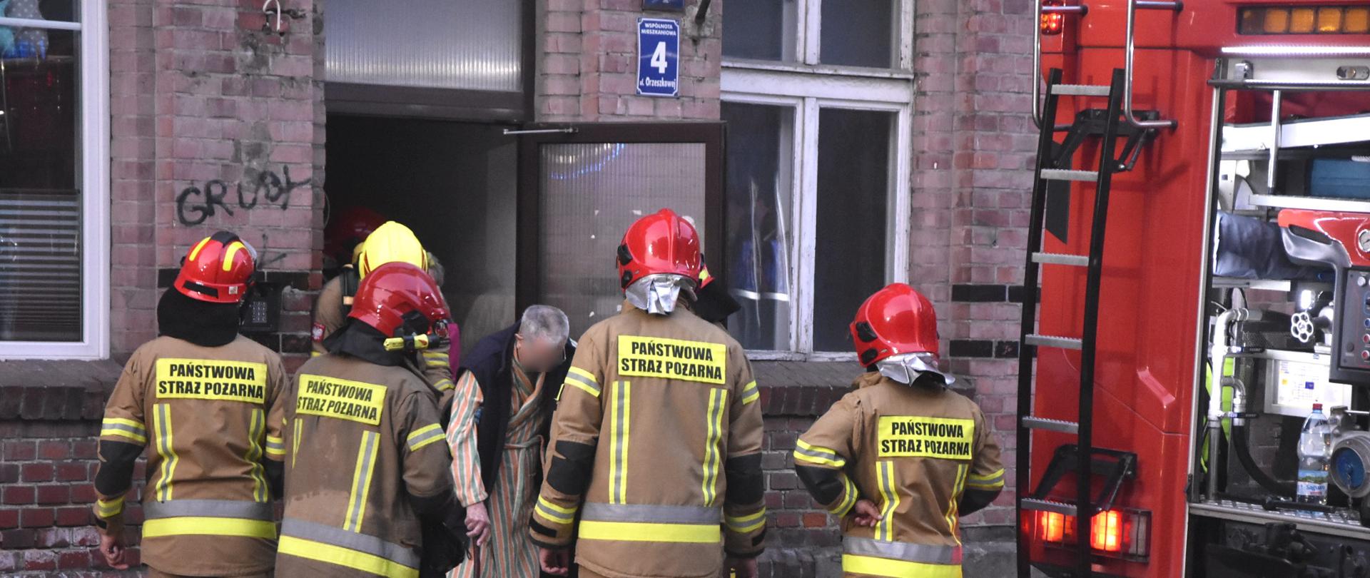 Fotografia przedstawia strażaków oraz osobę ewakuowaną z pomieszczenia budynku, w którym wybuchł pożar piwnicy
