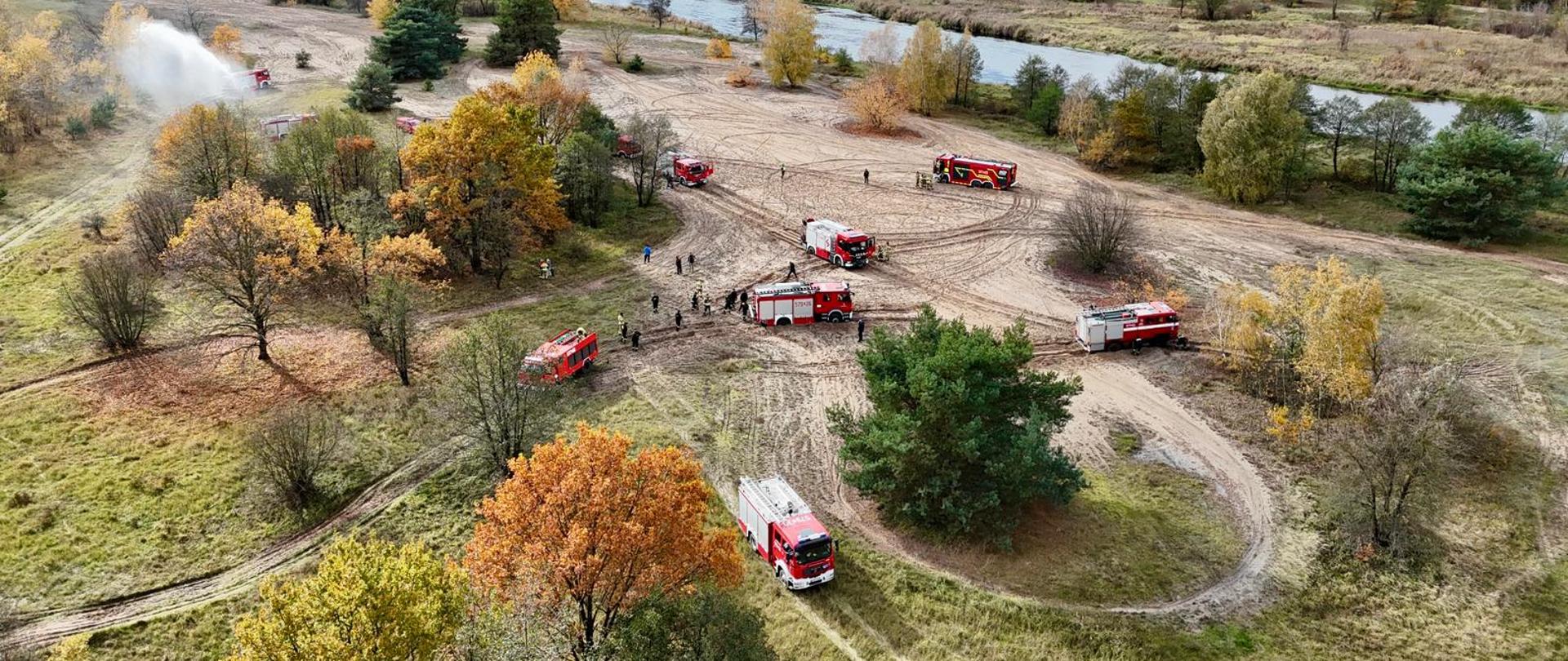 Z lotu ptaka widać kilka wozów strażackich rozmieszczonych na piaszczystym terenie w pobliżu rzeki. Strażacy prowadzą ćwiczenia z doskonalenia jazdy w trudnym terenie. Wokół jesienne drzewa w odcieniach żółci i pomarańczu.
