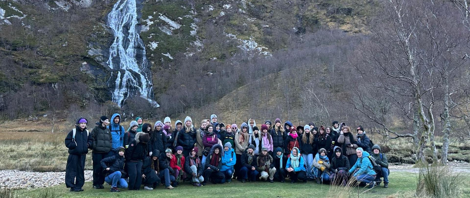 Na zdjęciu widzimy uczniów i nauczyciele biorący udział w plenerze. Fotografia została wykonana w plenerze. W tle widzimy wysokie zbocza gór z wodospadem. Steall Falls to wodospad o wysokości 120 m położony niedaleko Fort William w Szkocji, drugi co do wysokości w Wielkiej Brytanii. Położony jest w dolinie Glen Nevis.