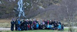 Na zdjęciu widzimy uczniów i nauczyciele biorący udział w plenerze. Fotografia została wykonana w plenerze. W tle widzimy wysokie zbocza gór z wodospadem. Steall Falls to wodospad o wysokości 120 m położony niedaleko Fort William w Szkocji, drugi co do wysokości w Wielkiej Brytanii. Położony jest w dolinie Glen Nevis.