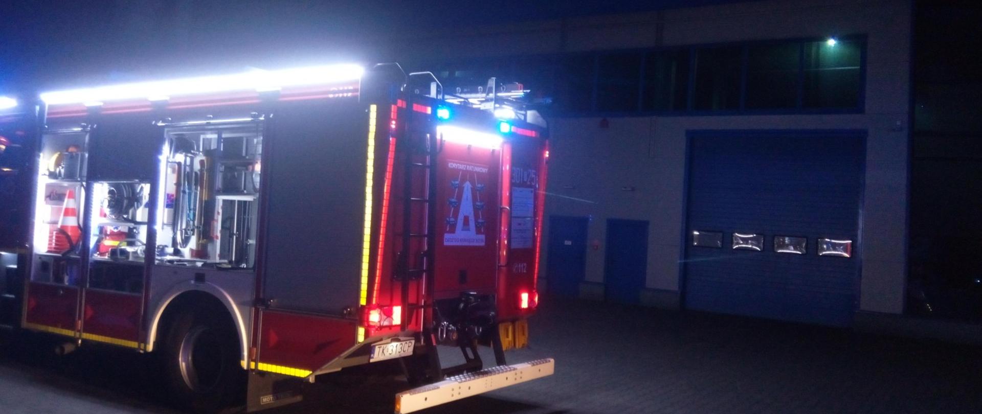 Zdjęcie przedstawia halę przemysłową na tle której stoi zaparkowany wóz strażacki.