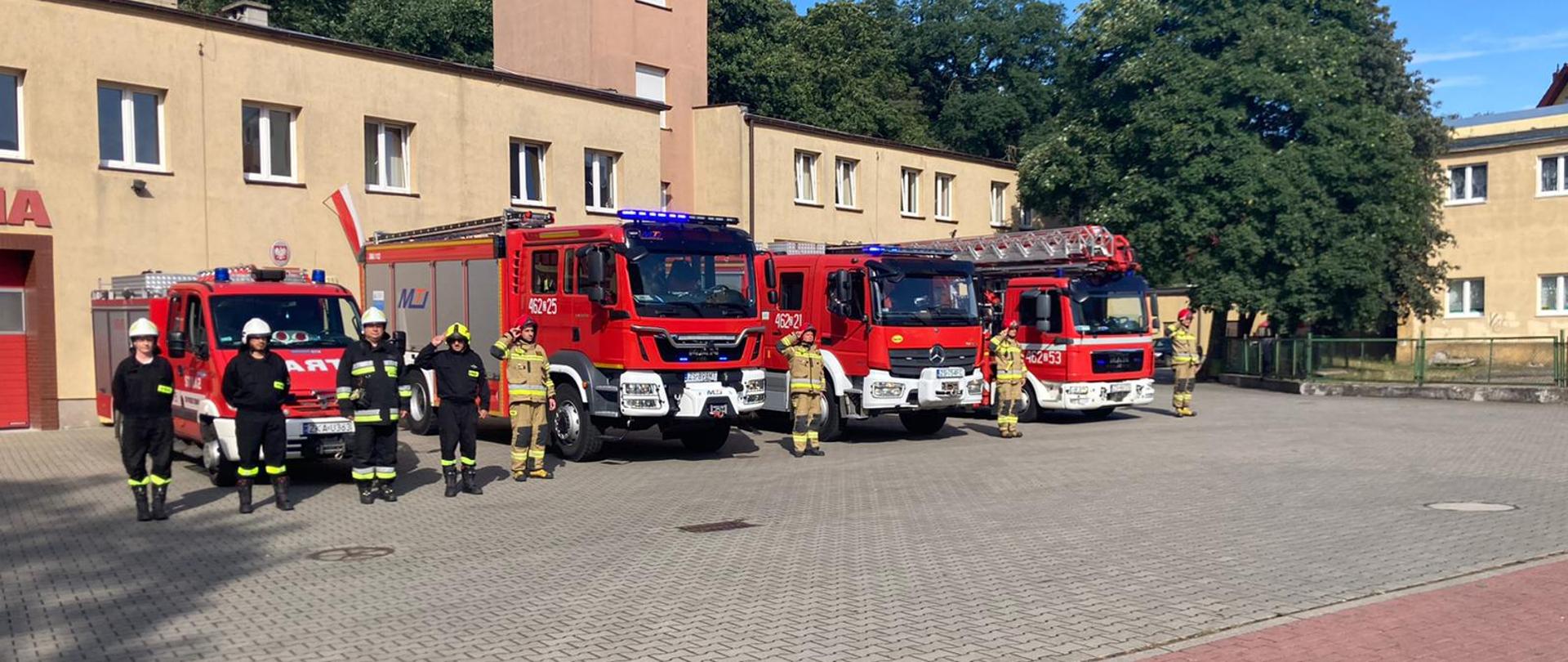 Zdjęcie przedstawia strażaków stojących na tle pojazdów straży pożarnej i remizy.