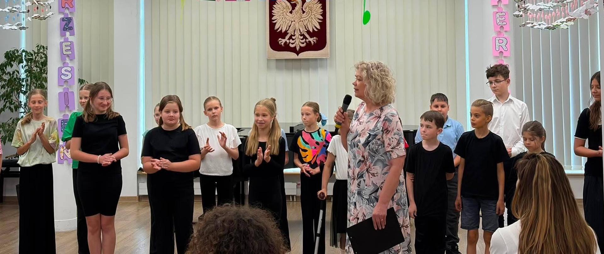 W sali koncertowej stoi grupa dzieci oraz kobieta. Kobieta przemawia przez mikrofon, który trzyma w ręku. Nad nimi podwieszone pod sufitem papierowe, kolorowe nuty. W tle na ścianie godło Polski.