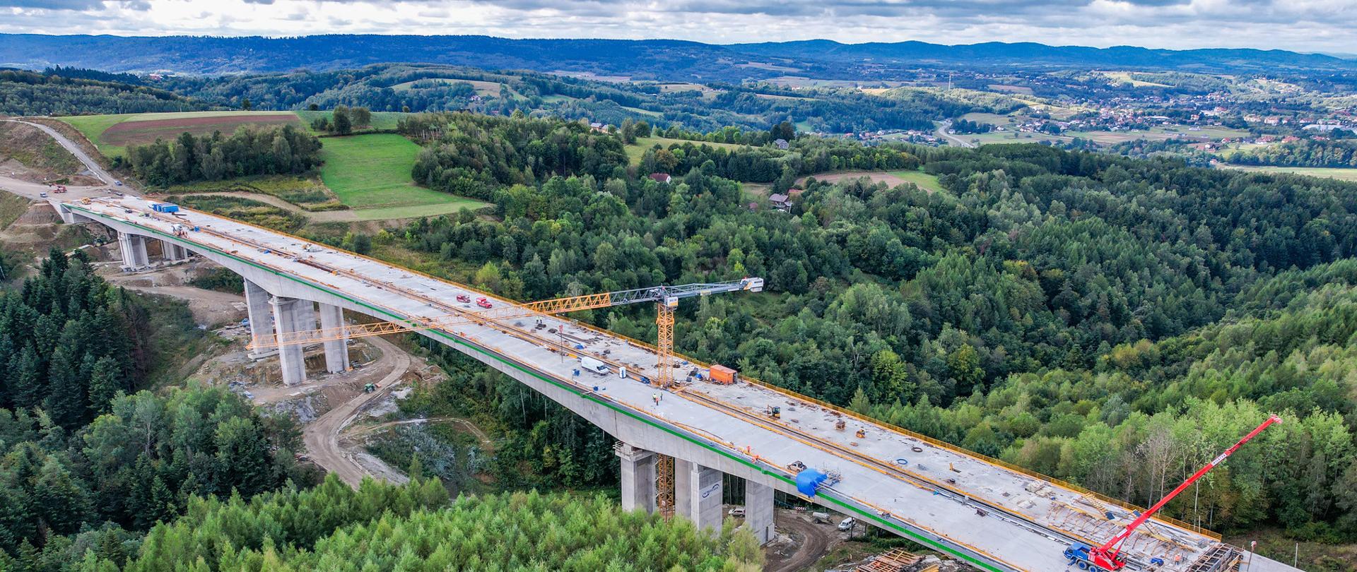 Budowana estakada ES-24 widziana z lotu ptaka. Betonowy obiekt na wysokich filarach przebiega nad zalesioną doliną. Na estakadzie pracują maszyny budowlane, a w tle rozciągają się pola, lasy i wzgórza.