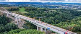 Budowana estakada ES-24 widziana z lotu ptaka. Betonowy obiekt na wysokich filarach przebiega nad zalesioną doliną. Na estakadzie pracują maszyny budowlane, a w tle rozciągają się pola, lasy i wzgórza.