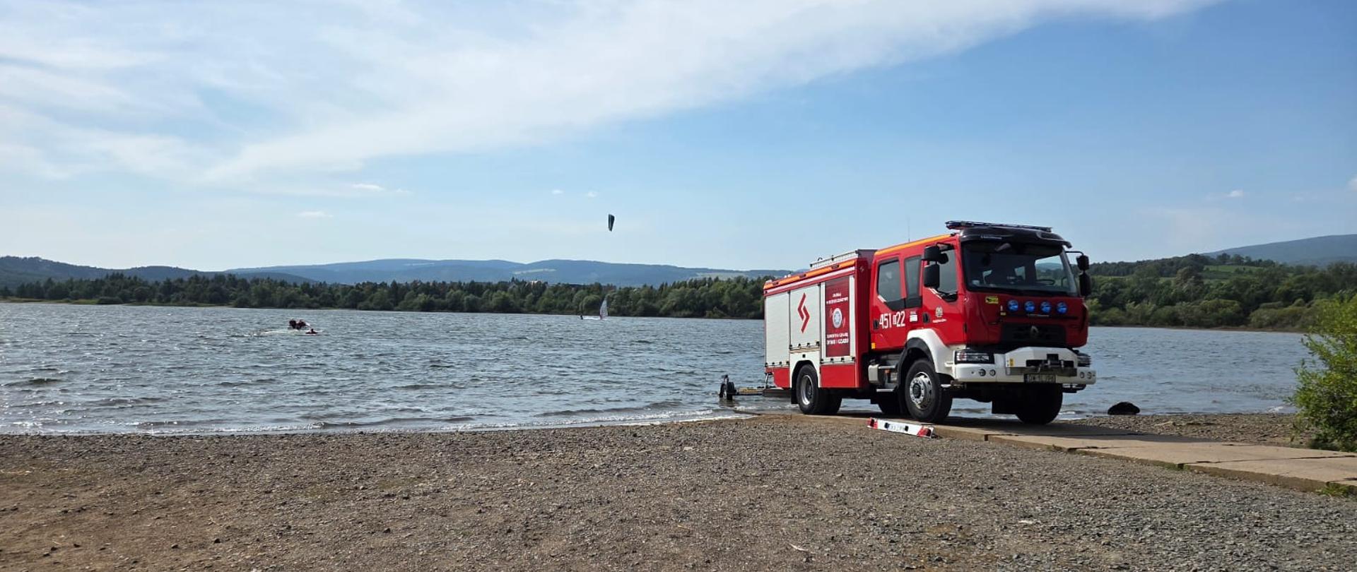 Pojazd ratowniczo gaśniczy ustawiony przy brzegu zbiornika wodnego w tle na wodzie dostrzegamy łódź z ratownikami.