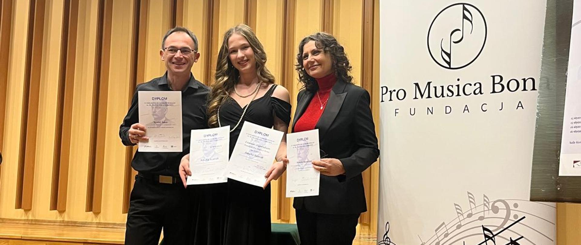 Na zdjęciu - Maciej Janas, Amelia_Szmidt, Monika_Brewczak. W tle scena konkursu i baner fundacji.