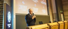 Zdjęcie z boku, wiceminister Gzik stoi i mów do trzymanego w ręku mikrofonu, za nim wbudowany w ścianę wielki ekran.