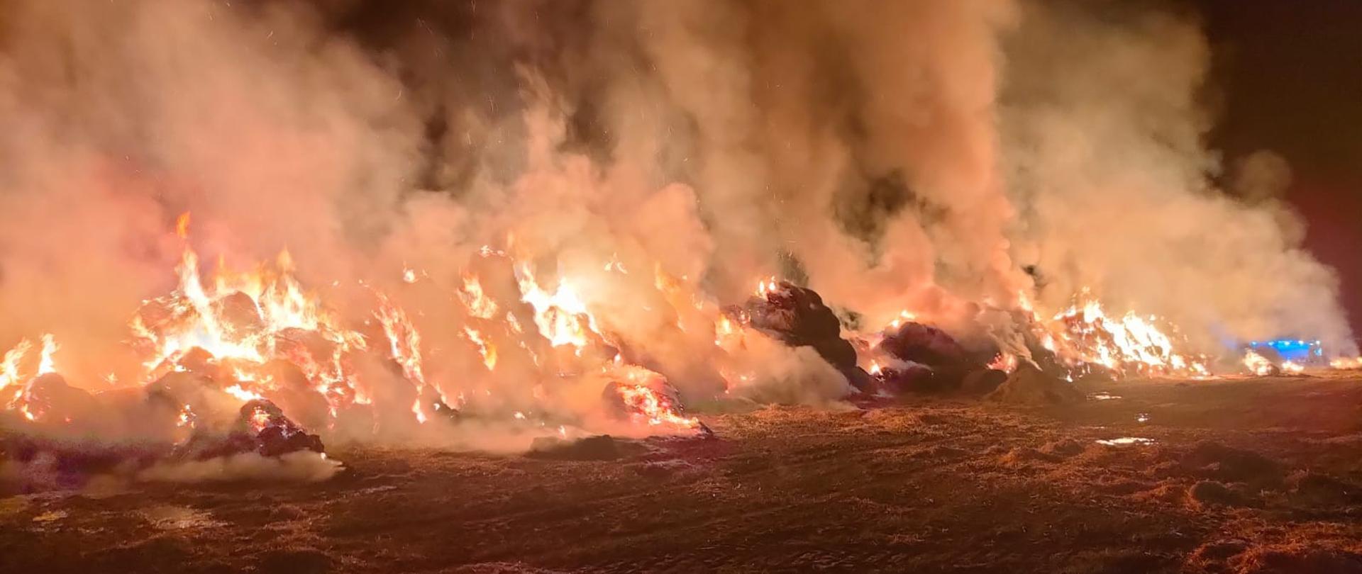 Na zdjęciu widać stertę słomy całkowicie objętą pożarem. W oddali za dymem widać niebieskie światła samochodu strażackiego. Jest ciemno.