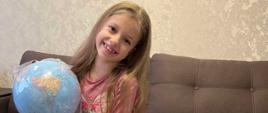 Do zdjęcia pozuje uśmiechnięta dziewczynka z globusem w rękach; przed dziewczynką leżą kolorowe przybory szkolne