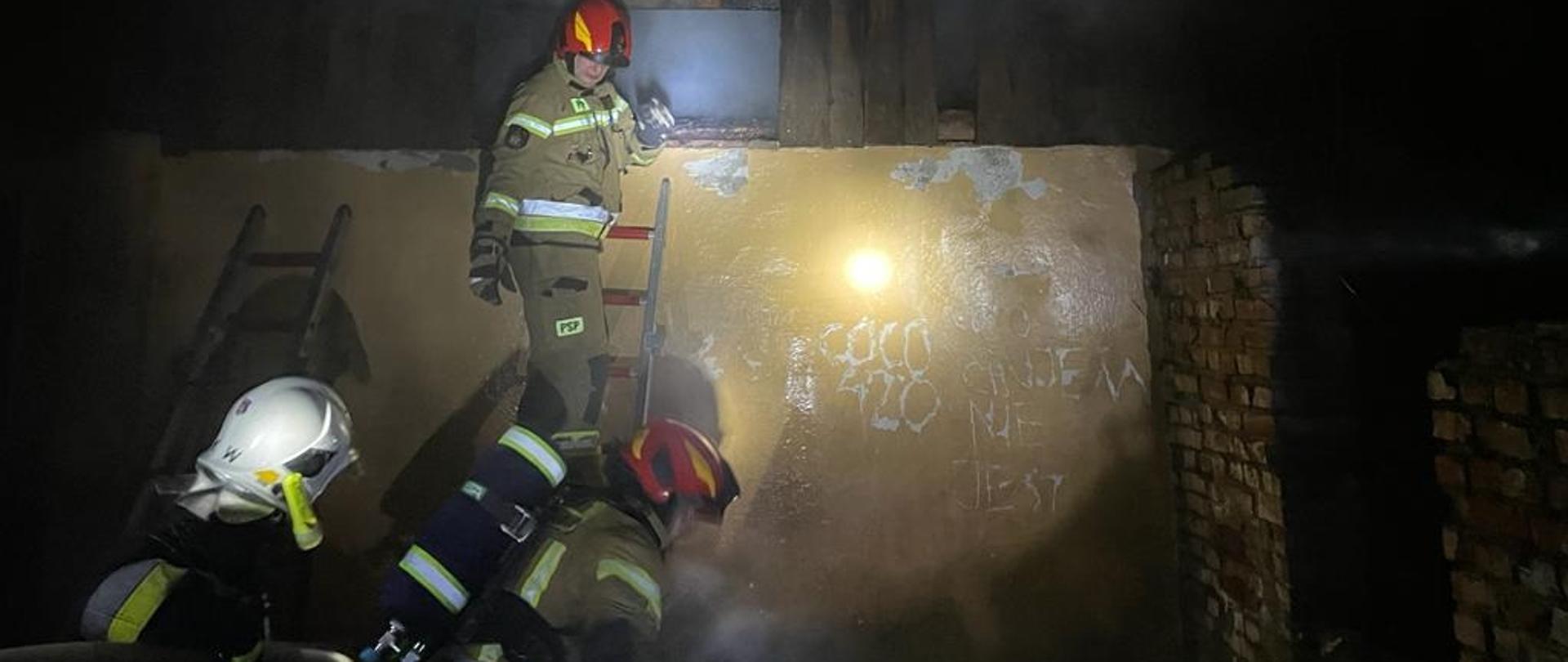 Zdjęcie zrobione nocą. Na ścianie budynku oparta drabina, na drabinie strażak w mundurze bojowym i czerwonym hełmie. Na dole jeszcze dwóch strażaków.