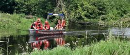 Fotografia przedstawia czterech ratowników siedzących w łodzi płaskodennej aluminiowej, która płynie po rzece, dwóch z nich ma ubrania do pracy w wodzie barwy czerwonej, szelki ratownicze oraz czerwone kaski na głowach, dwóch kamizelki barwy czerwonej z szelkami a jeden z nich ma kask na głowie barwy czerwonej a drugi czapkę z daszkiem. Jeden ratownik siedzi na rufie łodzi i obsługuje silnik motorowy, trzech siedzi na dziobie skierowani twarzami do siebie. Łódź ma zamontowaną belkę ze światłem alarmowym barwy niebieskiej, na jej boku widnieje napis PAŃSTWOWA STRAŻ POŻARNA oraz numer boczny 451EŁ2. Koryto rzeki zarośnięte trawą, dookoła drzewa, świeci słońce.