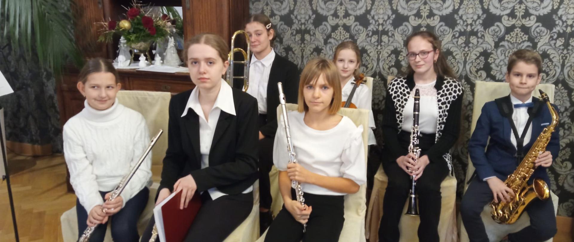 Zdjęcie przedstawia grupę uczniów siedzących na krzesłach, trzymających instrumenty. W tle ściana z ciemną wzorzystą tapetą , po prawej stronie stoi drewniany zabytkowy kredens ze świąteczną dekoracją