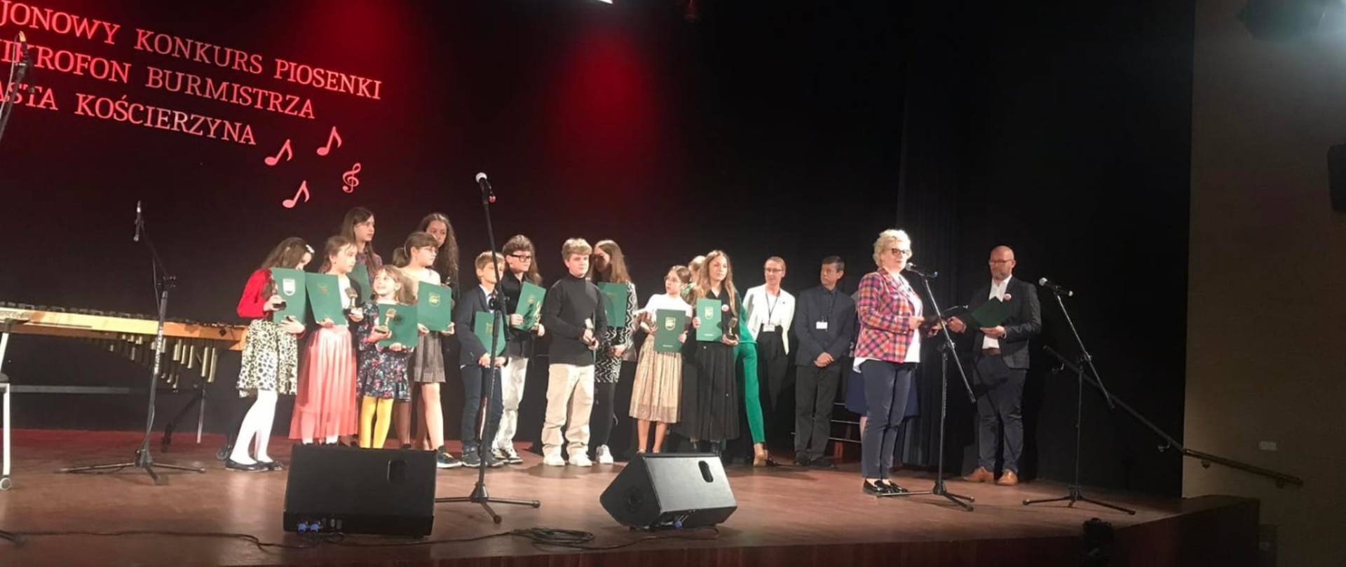 Na obrazku grupa dzieci w różnym wieku, którzy trzymają w rękach zielone teczki. Przy mikrofonie stoi kobieta- blondynka w ciemnych spodniach i kraciastej kolorowej marynarce.