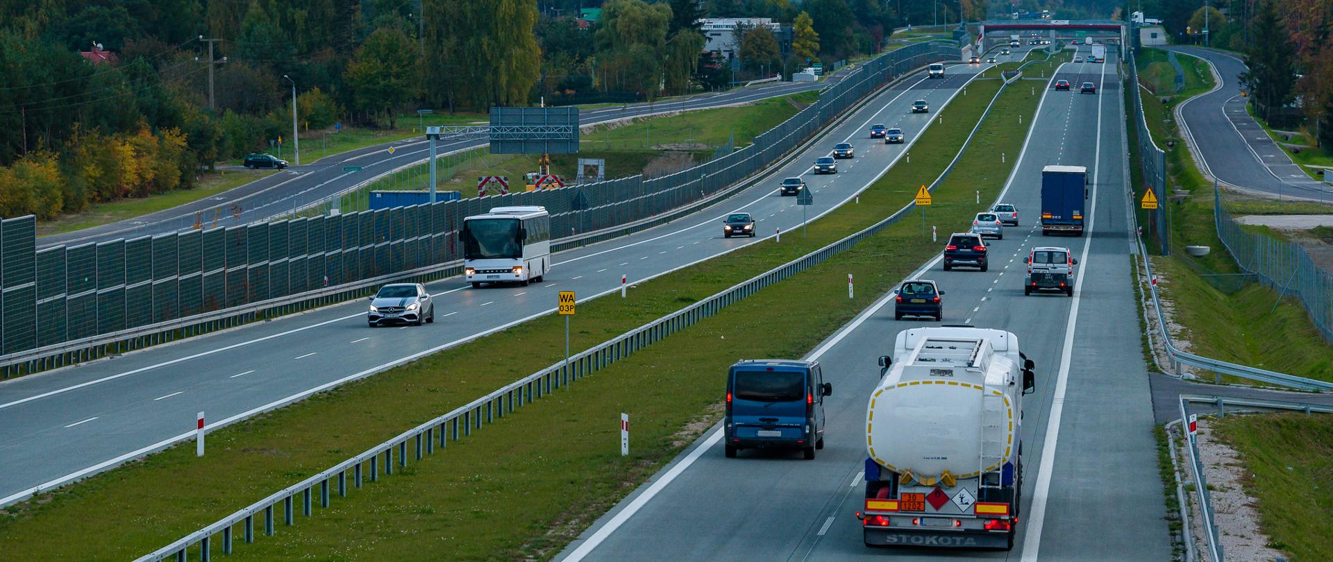 Dwujezdniowa droga ekspresowa z intensywnym ruchem pojazdów. Na jezdniach poruszają się samochody osobowe, autobus i samochód ciężarowy, a trasę otaczają ekrany akustyczne, bariery ochronne oraz tereny zielone.