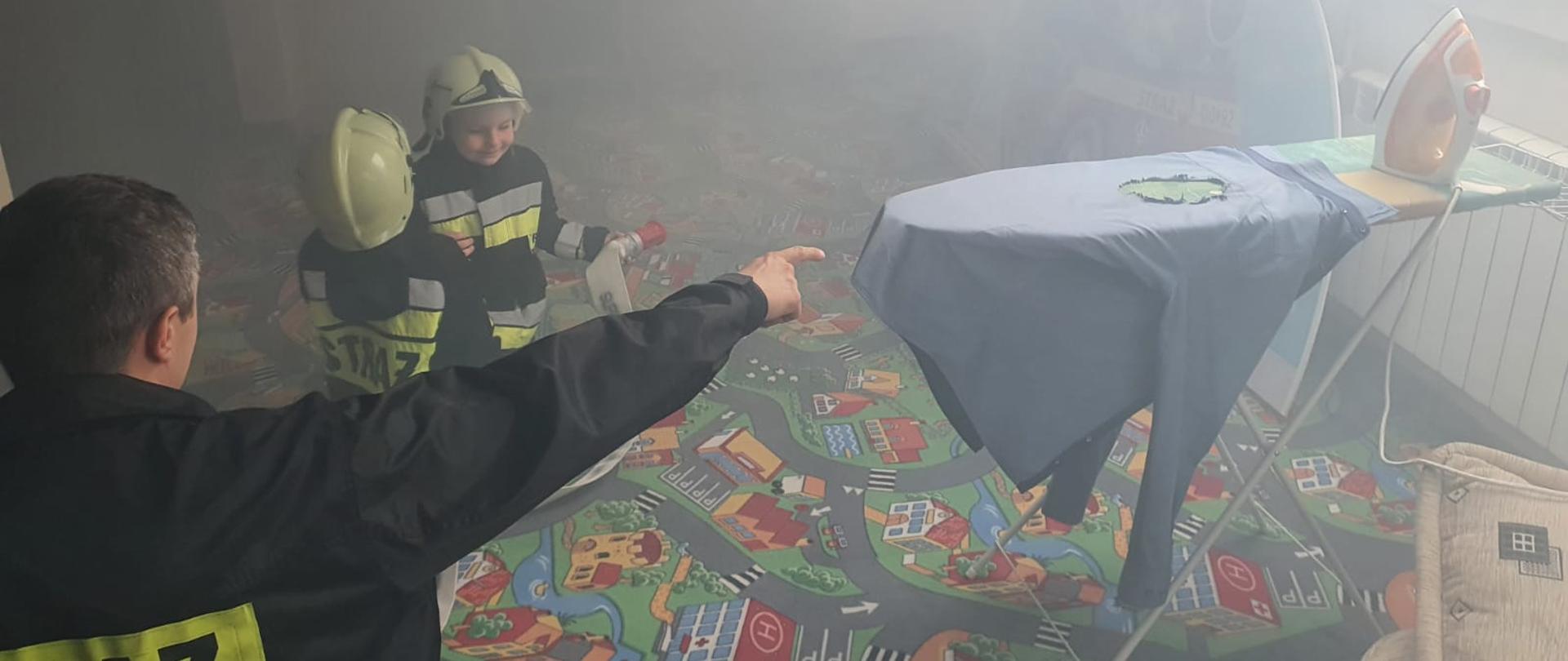Zdjęcie przedstawia strażaka, który gestem ręki wskazuje na prasowalnicę, na której leży koszula z wypalona dziurą. Dwójka małych dzieci trzyma w rękach prądownice wodną oraz wąż strażacki. Wszyscy znajdują się na sali edukacyjnej Ognik, w której panuje niewielkie zadymienie.