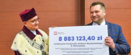 Minister Czarnek i mężczyzna w czerwonym ceremonialnym stroju stoją obok siebie i trzymają wielki symboliczny czek z napisem 8 883 123,40 zł.