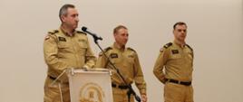 Scena Auli SA PSP w Krakowie. Trzech mężczyzn, strażacy w umundurowaniu służbowym. Przy mównicy mł.bryg. Dariusz Ruchała za nim komendant – nadbryg. Piotr Filipek oraz p.o. zastępca komendanta mł.bryg. Bogusław Szydło