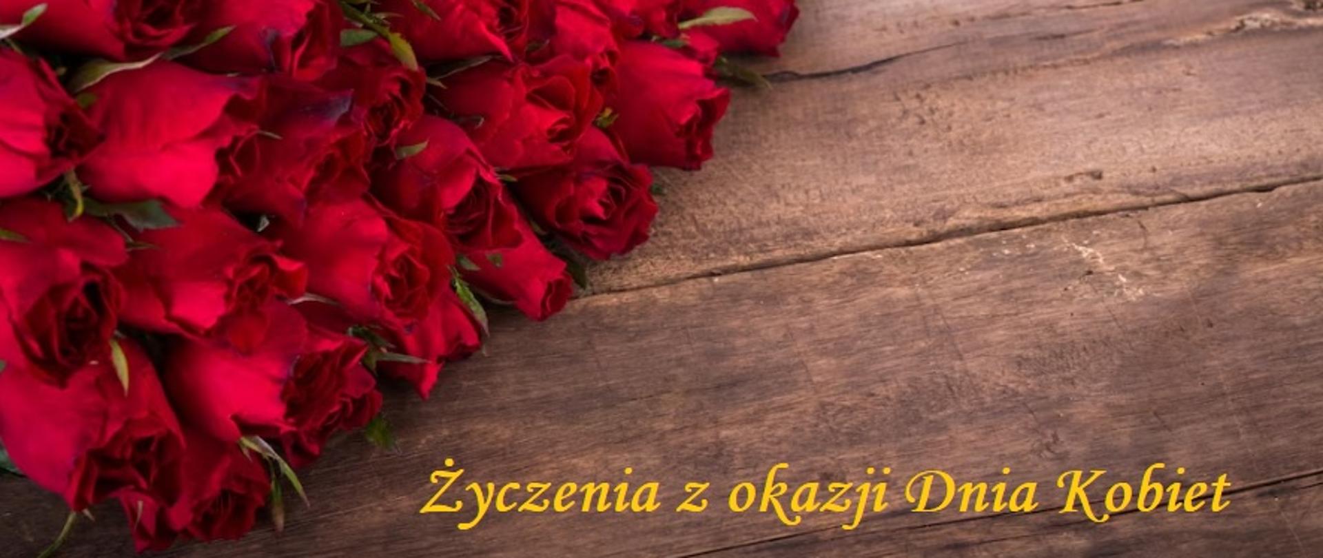 Czerwone róże na leżą po lewej stronie na drewnianych deskach. Na dole z prawej strony żółty napis Życzenia z okazji Dnia Kobiet