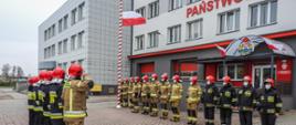 Zdjęcie przedstawia uroczystą zmianę służby w dniu 2 maja 2021 r. Podczas zmiany służby przed budynkiem komendy podniesiono flagę Polski i odegrano hymn państwowy. Strażacy występujący podczas zmiany ubrani byli w ubrania specjalne.