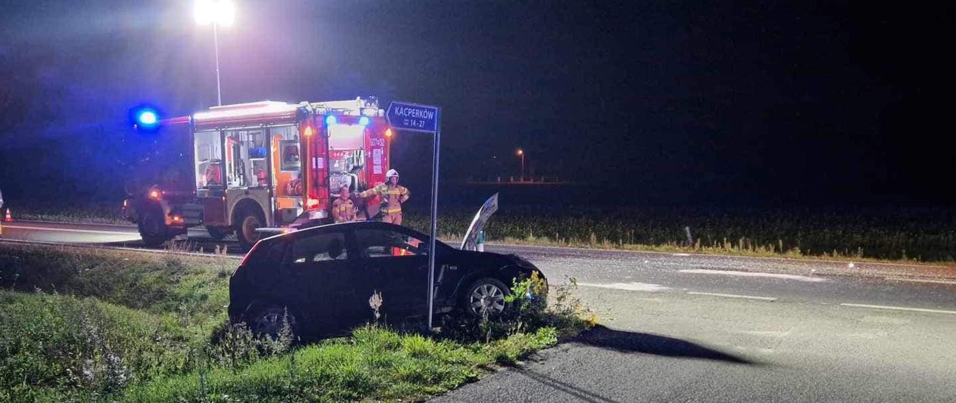 Nocna scena przedstawia skutki wypadku samochodowego na skrzyżowaniu drogi krajowej nr 48 z drogą gminną. Ciemny hatchback leży w rowie przy drodze, z uszkodzonym przodem i uchyloną maską. W tle widać zaparkowany na drodze czerwony wóz strażacki z migającymi niebieskimi światłami, a obok niego dwóch strażaków w odzieży ochronnej. Obok uszkodzonego samochodu widoczny jest znak drogowy z napisem „KACPERKÓW” i kilkoma numerami. Teren jest oświetlony światłami pojazdów ratunkowych i latarnią uliczną. Droga miejscami wydaje się podmokła, a wzdłuż jej krawędzi porasta ją rzadka roślinność.