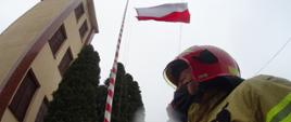 Na zdjęciu po prawej stronie strażak w ubraniu specjalnym oddaje honory podczas wciągania flagi na maszt. Po lewej stronie budynek komendy i drzewa.