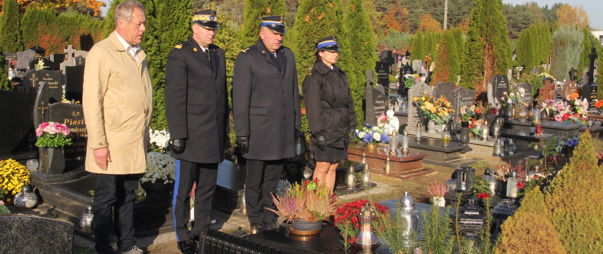 strażacy oraz mężczyzna w jasnym płaszczu stoją przed grobem na cmentarzu
