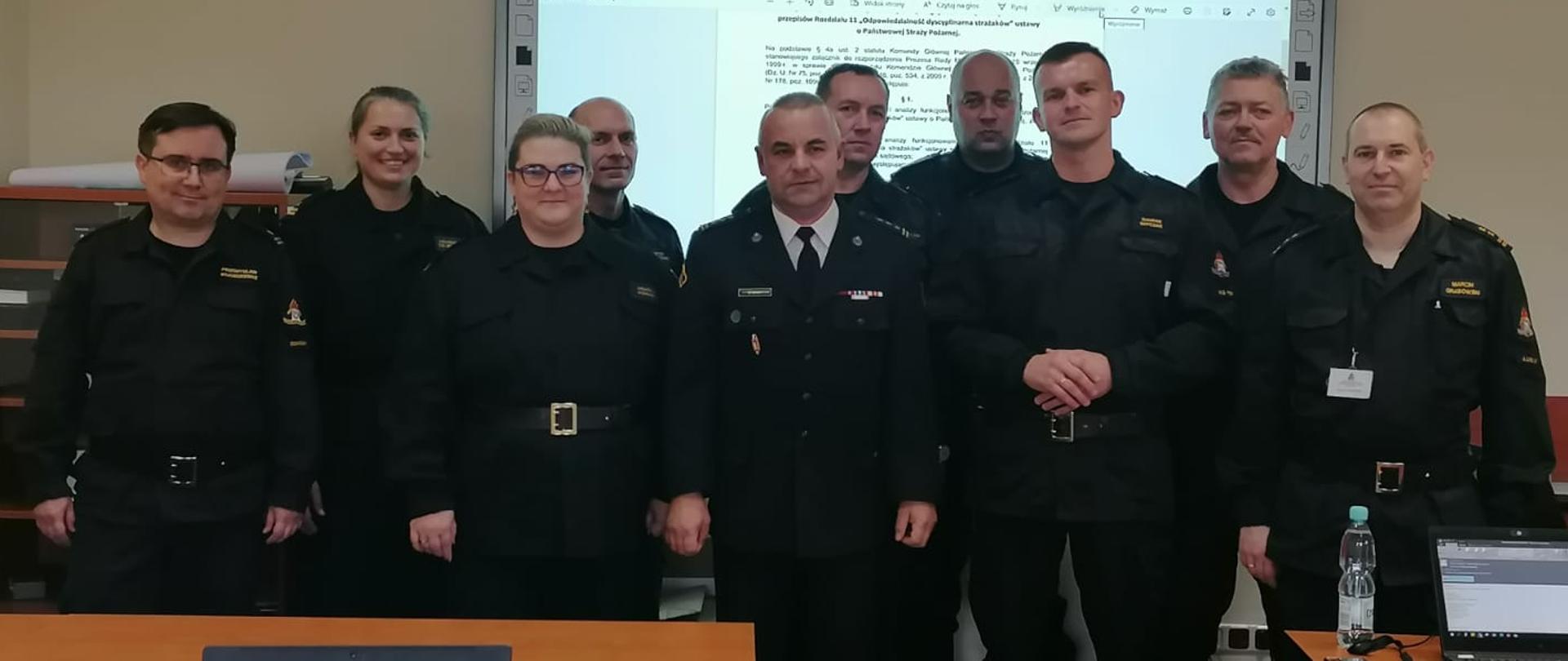 Na zdjęciu widać dziesięciu strażaków podczas spotkania roboczego w sali wykładowej. Strażacy ustawieni są do zdjęcia grupowego.