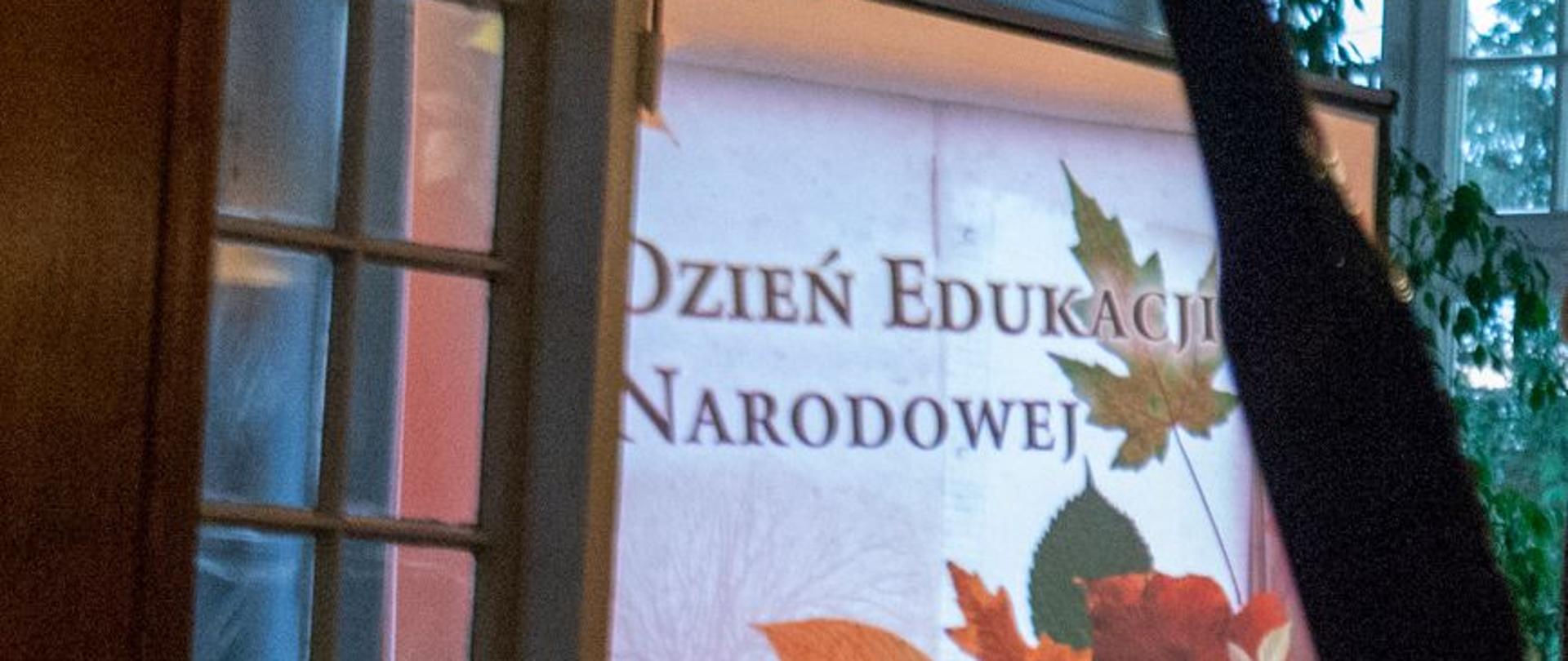 Zdjęcie kolorowe. Ekran rzutnika z wyświetlonym kolorowym napisem Dzień Edukacji Narodowej. W tle kolorowe liście.