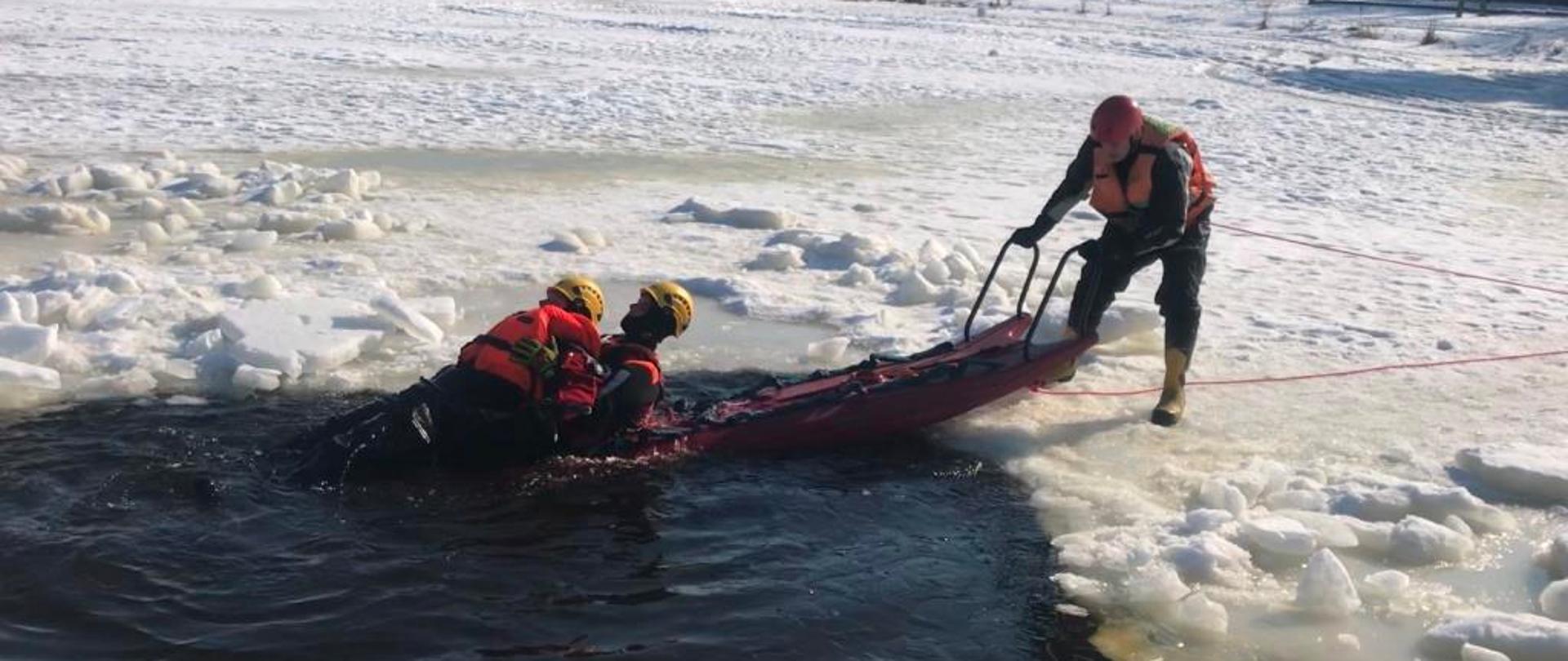 Zdjęcie przedstawia trzech strażaków ubranych w specjalne skafandry do działań na akwenach wodnych. Dwaj z nich znajdują się w wodzie. Pierwszy udaje osobę tonącą pod którą załamał się lód. Drugi próbuje go uratować. Trzeci z nich trzyma specjalne sanie przystosowanych do poruszania się po lodzie i prowadzeniu działań ratowniczych. Ratownik numer dwa podejmuje próbę wydobycia osoby poszkodowanej na sanie. Dookoła nich znajduje się śnieg. 