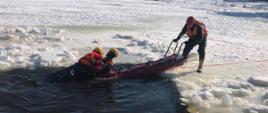 Zdjęcie przedstawia trzech strażaków ubranych w specjalne skafandry do działań na akwenach wodnych. Dwaj z nich znajdują się w wodzie. Pierwszy udaje osobę tonącą pod którą załamał się lód. Drugi próbuje go uratować. Trzeci z nich trzyma specjalne sanie przystosowanych do poruszania się po lodzie i prowadzeniu działań ratowniczych. Ratownik numer dwa podejmuje próbę wydobycia osoby poszkodowanej na sanie. Dookoła nich znajduje się śnieg.