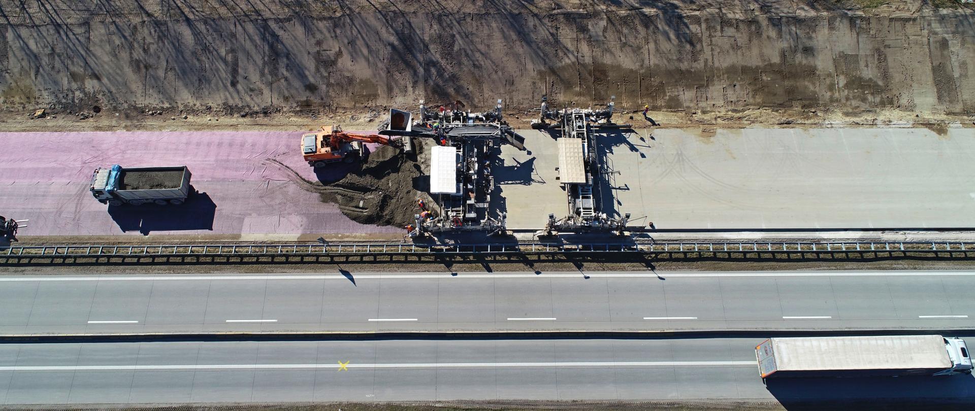 Układanie górnych warstw nawierzchni betonowej. Widoczna pracująca maszyna rozkładająca masę na rozłożoną agrowłókninę, stanowiącą jedną z warstw.