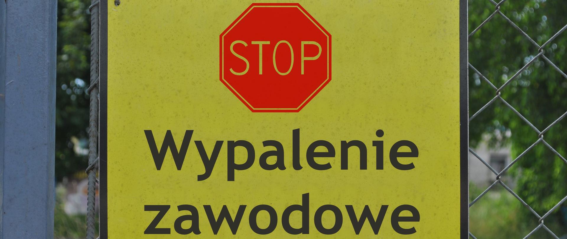 Na ogrodzeniu wisi żółta tablica, na której znajduje się czerwony znak „STOP" a pod nim czarny napis „Wypalenie zawodowe"