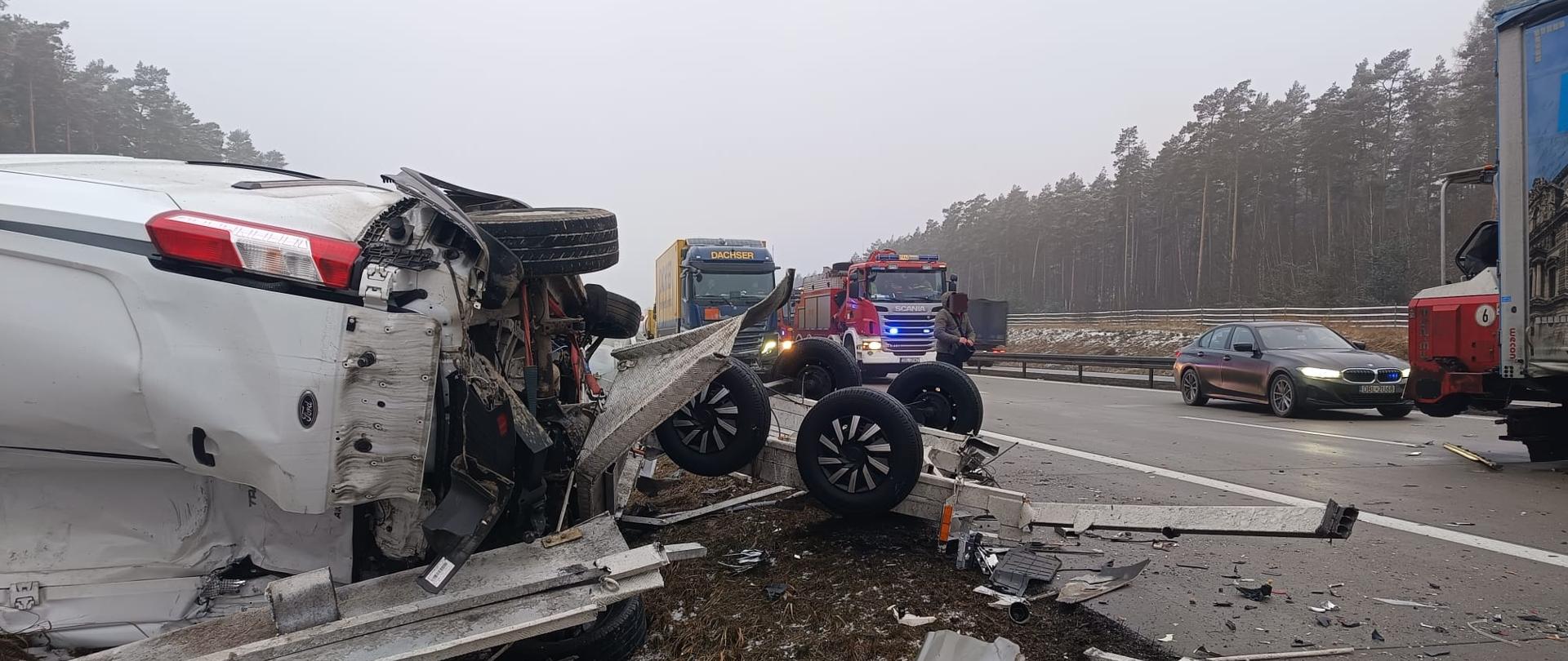 
Zdjęcie przedstawia miejsce groźnego wypadku drogowego na trasie szybkiego ruchu w ponury, mglisty dzień. Na pierwszym planie po lewej stronie widoczny jest mocno rozbity biały samochód dostawczy marki Ford, który leży przewrócony na boku. Obok niego znajduje się uszkodzona, wywrócona kołami do góry dwuosiowa przyczepa, z której na jezdnię i pobocze rozsypały się odłamki karoserii. W tle, na zablokowanych pasach drogi, stoją samochody ciężarowe (w tym jeden z napisem Dachser) oraz wóz straży pożarnej na sygnale. Po prawej stronie, na sąsiednim pasie, obok miejsca zdarzenia przejeżdża ciemne BMW. Wzdłuż drogi ciągnie się gęsty, zamglony las iglasty.