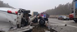
Zdjęcie przedstawia miejsce groźnego wypadku drogowego na trasie szybkiego ruchu w ponury, mglisty dzień. Na pierwszym planie po lewej stronie widoczny jest mocno rozbity biały samochód dostawczy marki Ford, który leży przewrócony na boku. Obok niego znajduje się uszkodzona, wywrócona kołami do góry dwuosiowa przyczepa, z której na jezdnię i pobocze rozsypały się odłamki karoserii. W tle, na zablokowanych pasach drogi, stoją samochody ciężarowe (w tym jeden z napisem Dachser) oraz wóz straży pożarnej na sygnale. Po prawej stronie, na sąsiednim pasie, obok miejsca zdarzenia przejeżdża ciemne BMW. Wzdłuż drogi ciągnie się gęsty, zamglony las iglasty.