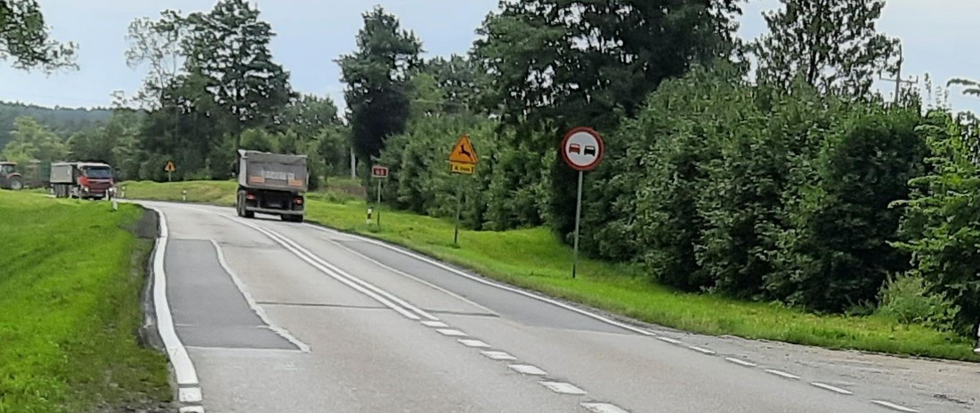Odcinek drogi wśród drzew i łąk. Nawierzchnia drogi jest zniszczona i pofałdowana. Na drodze samochody ciężarowe.