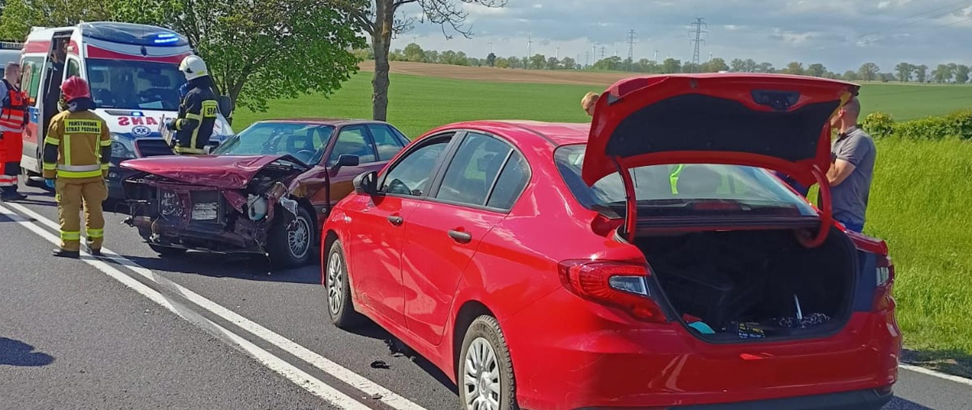 dwa rozbite samochody na lewym pasie drogi, dalej dwóch strażaków, ratownik medyczny, karetka