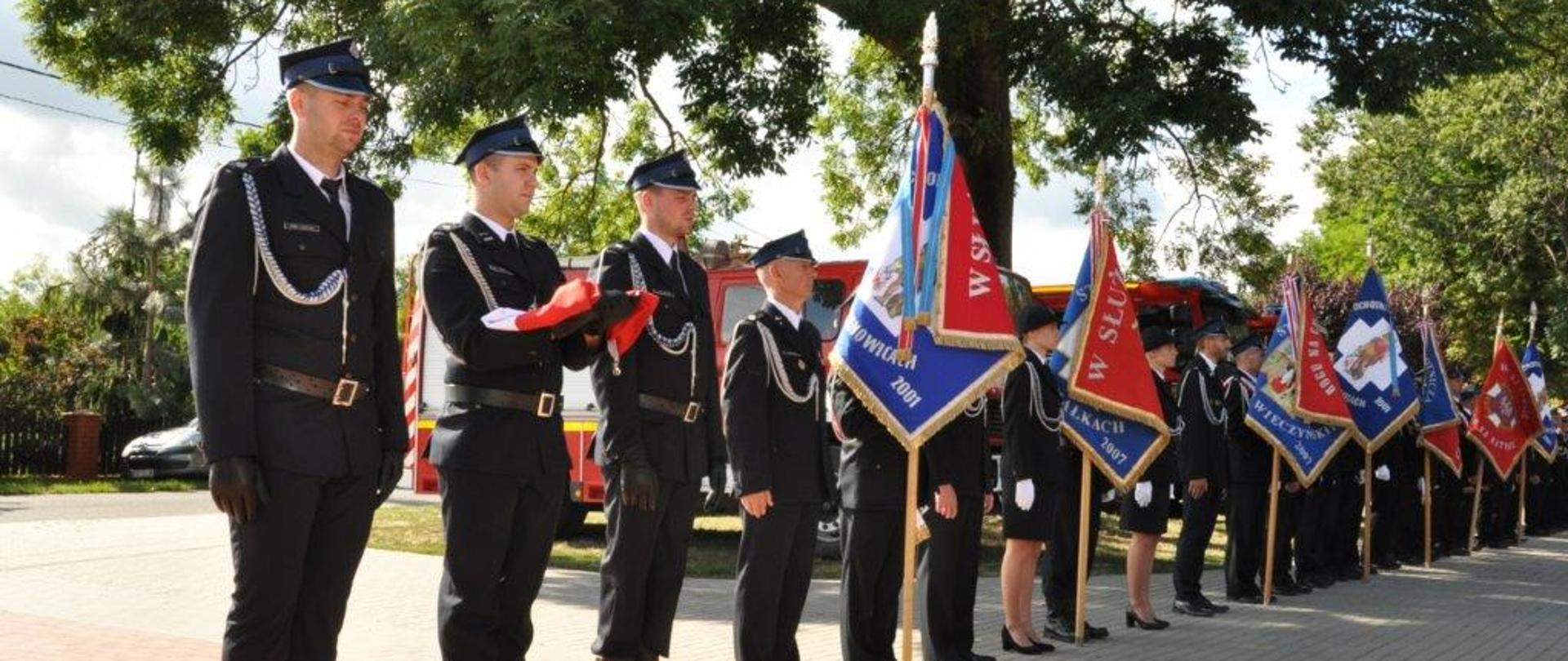 Szereg stojących na baczność osób ubranych w mundury OSP, część z nich trzyma sztandary, na pierwszym planie stoi t5rzech, z których środkowy trzyma złożoną flagę