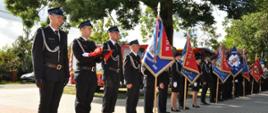 Szereg stojących na baczność osób ubranych w mundury OSP, część z nich trzyma sztandary, na pierwszym planie stoi t5rzech, z których środkowy trzyma złożoną flagę