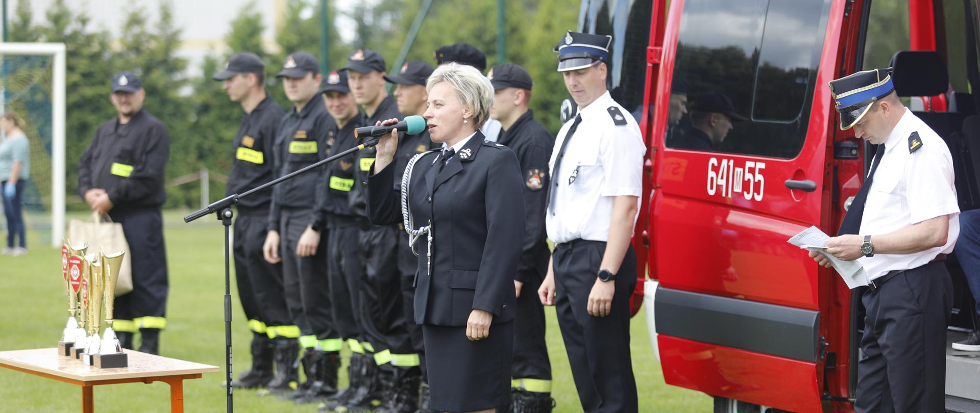Gminne Zawody Sportowo – Pożarnicze jednostek OSP z terenu gminy Repki na zdjęciu strażacy w ubraniu koszarowym stoją w szeregu na zbiórce na boisku trawiastym. po lewej stronie stoi czerwony bus strażacki