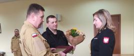 Komendant Powiatowy w Grodzisku Mazowieckim st. bryg. Krzysztof Tryniszewski wraz ze swoim zastępcą st. bryg. Piotrem Księżakiem wręczają kwiaty i pamiątkowy dyplom mł. kpt. Marioli Piątkowskiej odchodzącej na emeryturę. 