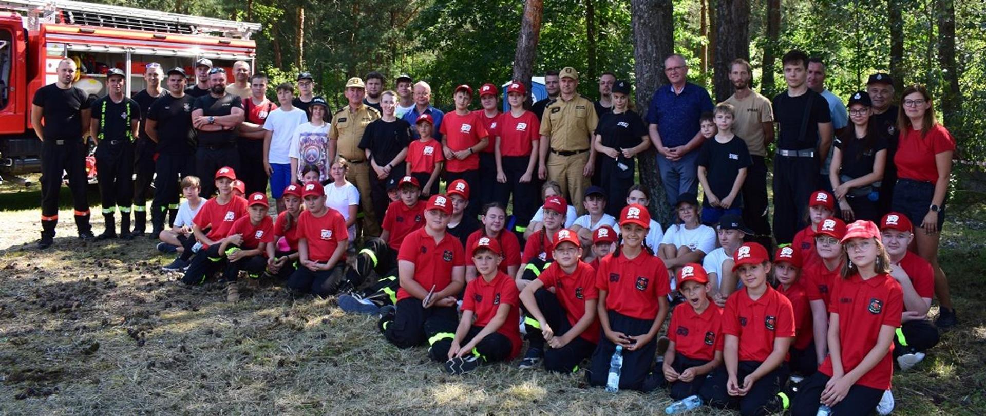 Fotografia wykonana podczas warsztatów Młodzieżowych Drużyn Pożarniczych w Wojkowie na terenie ośrodku wypoczynkowego Balaton. Na zdjęciu siedzą i stoją członkowie młodzieżowych drużyn pożarniczych z powiatu mieleckiego. Zdjęcie wykonane na tle drzew sosnowych. Między dziećmi na pamiątkowej fotografii stoją między innymi: poseł na Sejm RP Fryderyk Kapinos, st.bryg. Andrzej Marczenia zastępca podkarpackiego komendanta wojewódzkiego PSP oraz bryg. Łukasz Kapinos komendant powiatowy PSP w Mielcu. Z lewej strony zdjęcia widać czerwony samochód pożarniczy.