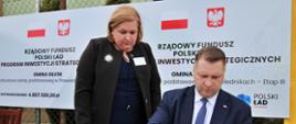 Minister Czarnek siedzi przy przykrytym zielonym materiałem stoliku, podpisuje dokumenty, nad nim stoi ubrana na czarno kobieta, za nimi ogrodzenie z siatki na którym wiszą plakaty z napisem Rządowy fundusz Polski Ład.